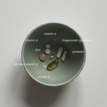 A minimalist photo of a green bowl containing various labeled supplements, including iron and vitamin B, omega-3, vitamin D, zinc, vitamin A, magnesium glycinate, and a probiotic—illustrating the complexity of modern supplement routines.
